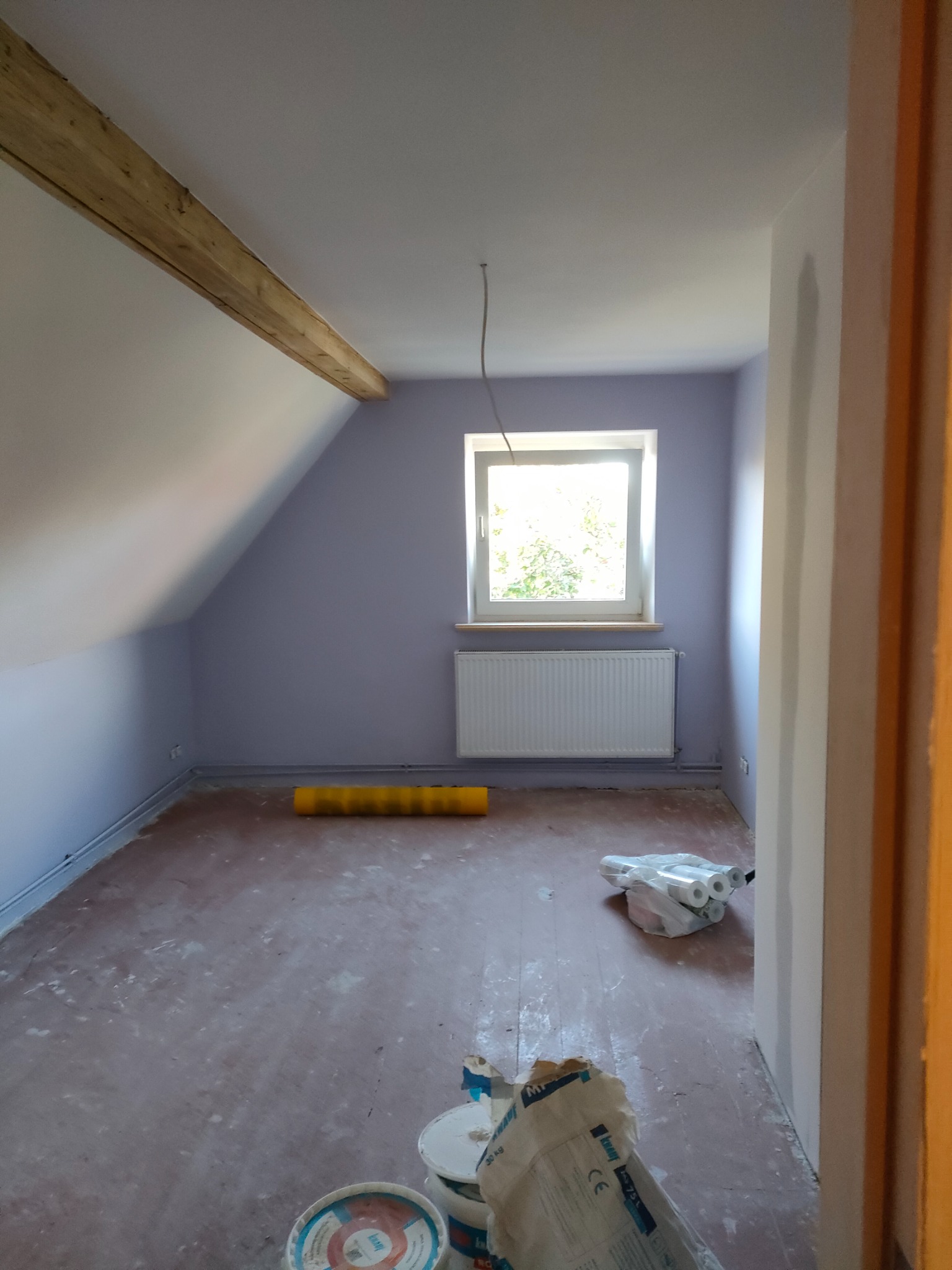 Poddasze po wstępnym malowaniu ścian na fioletowo, widoczna drewniana belka stropowa, okno z widokiem na zieleń, grzejnik pod oknem, żółty wałek malarski i worki z materiałami budowlanymi na podłodze.