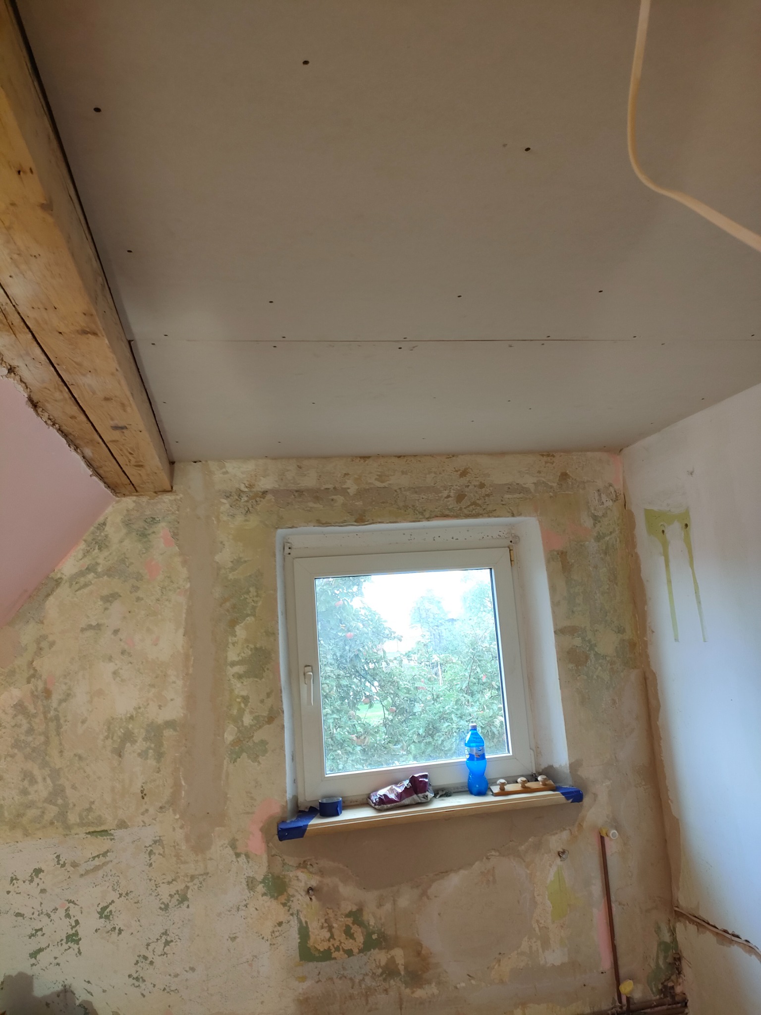 Pokój w trakcie prac przygotowawczych: ściany z resztkami starej farby i tapet, częściowo zakryte płytami kartonowo-gipsowymi na suficie, widoczne drewniane belki stropowe, okno z butelką wody...