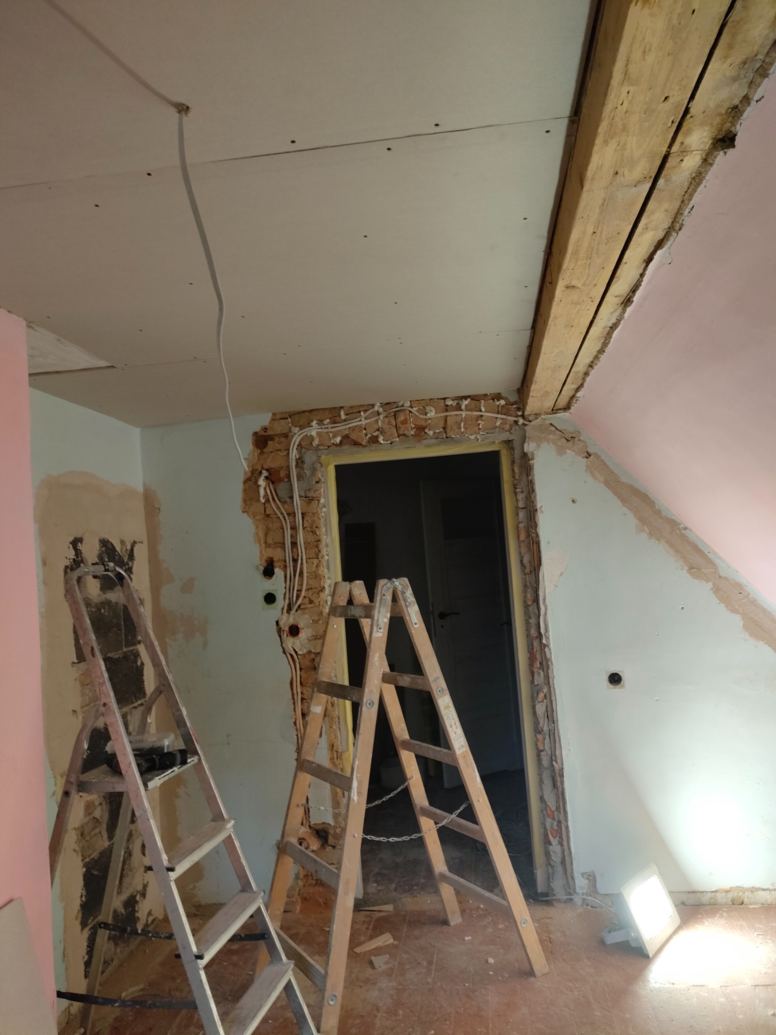 Pokój na poddaszu w trakcie prac: widoczne dwie drabiny, odsłonięta cegła wokół futryny drzwi, instalacja elektryczna, częściowo położona płyta gipsowo-kartonowa na suficie, drewniana belka...