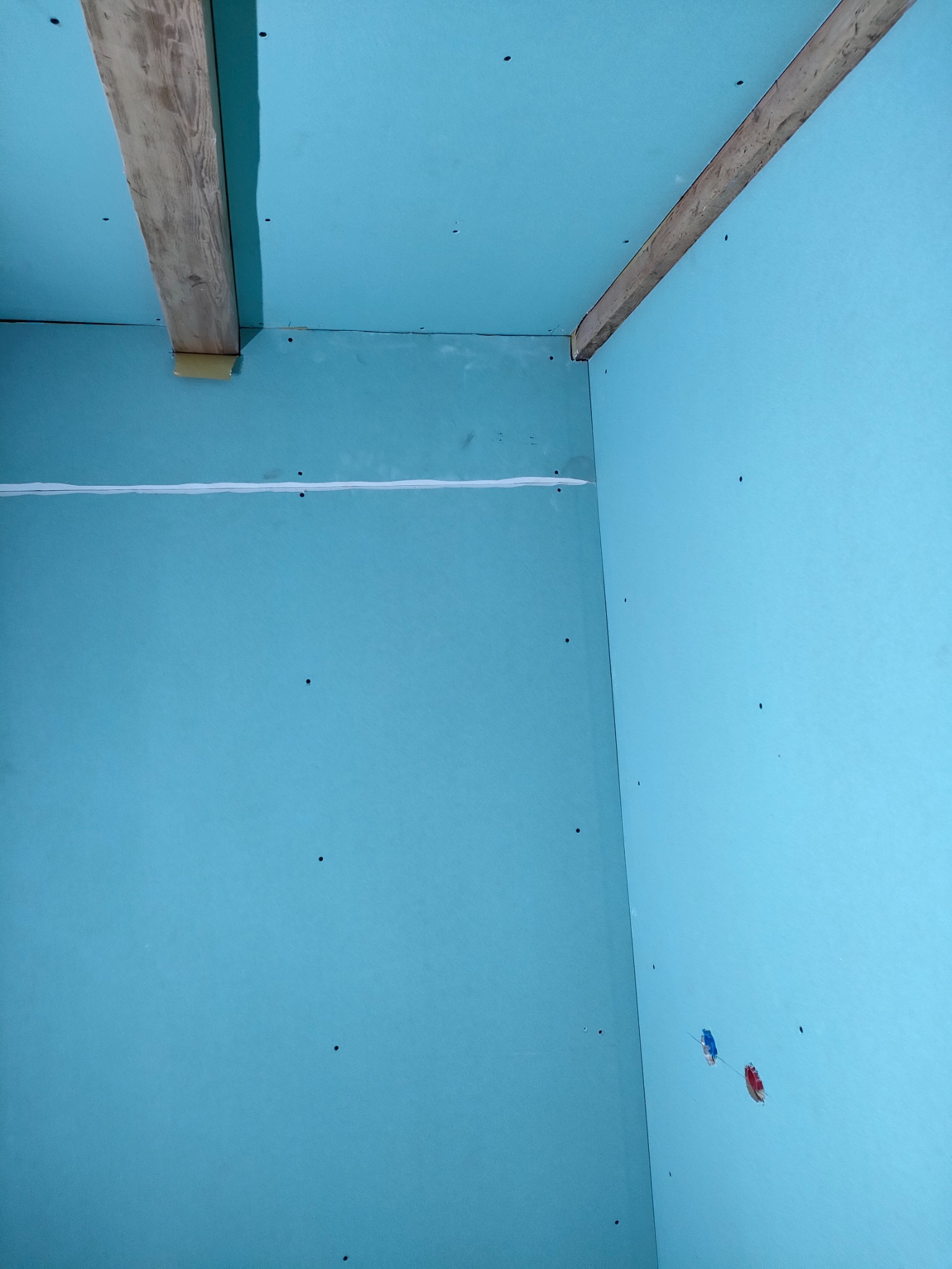 Niebieskie płyty gipsowo-kartonowe zamontowane na ścianach i suficie, widoczne drewniane belki konstrukcyjne i przewody elektryczne w narożniku pomieszczenia.