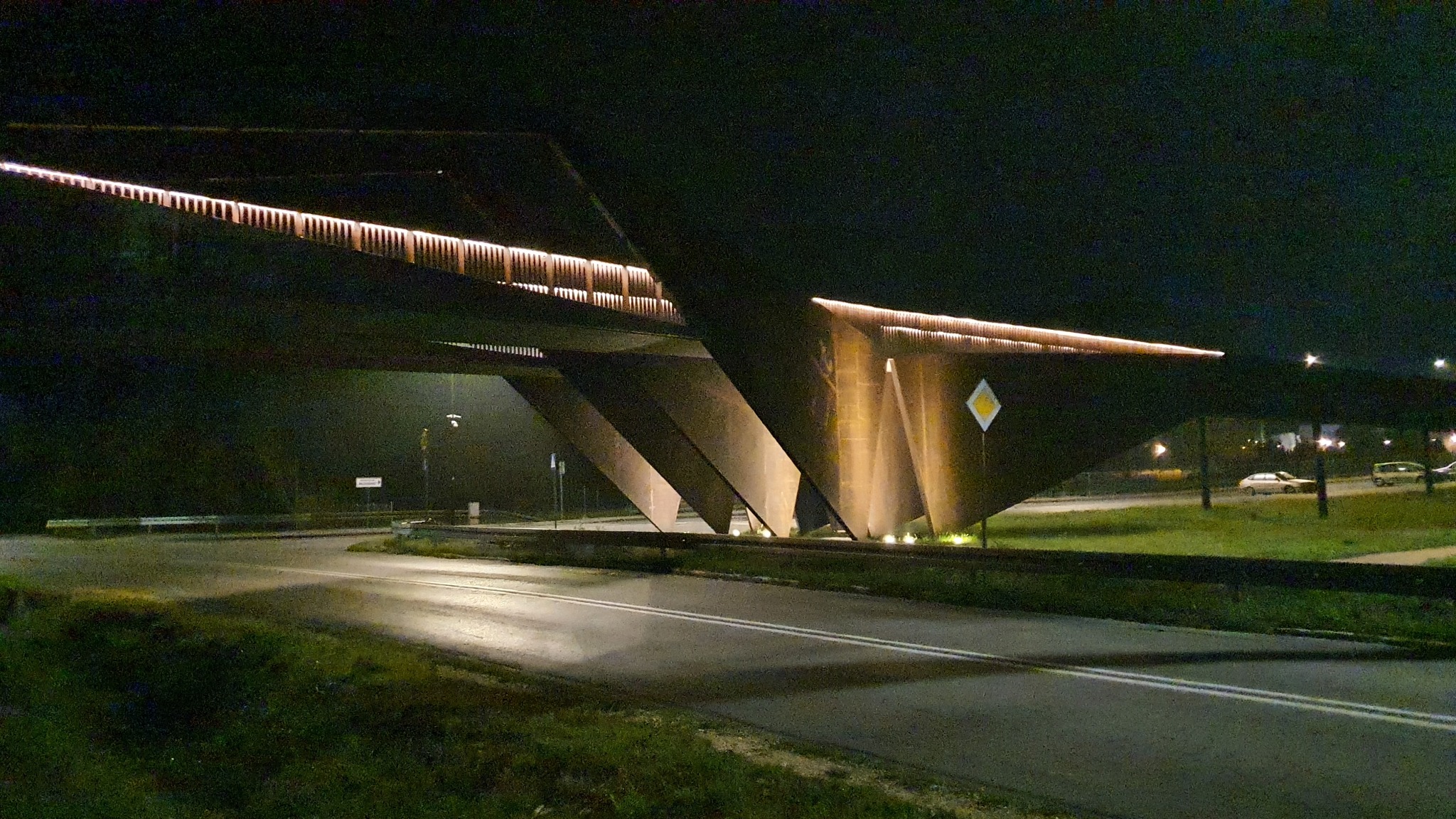 Nowoczesna iluminacja mostu w nocy, z widocznymi elementami oświetleniowymi LED i znakiem drogowym ostrzegawczym.