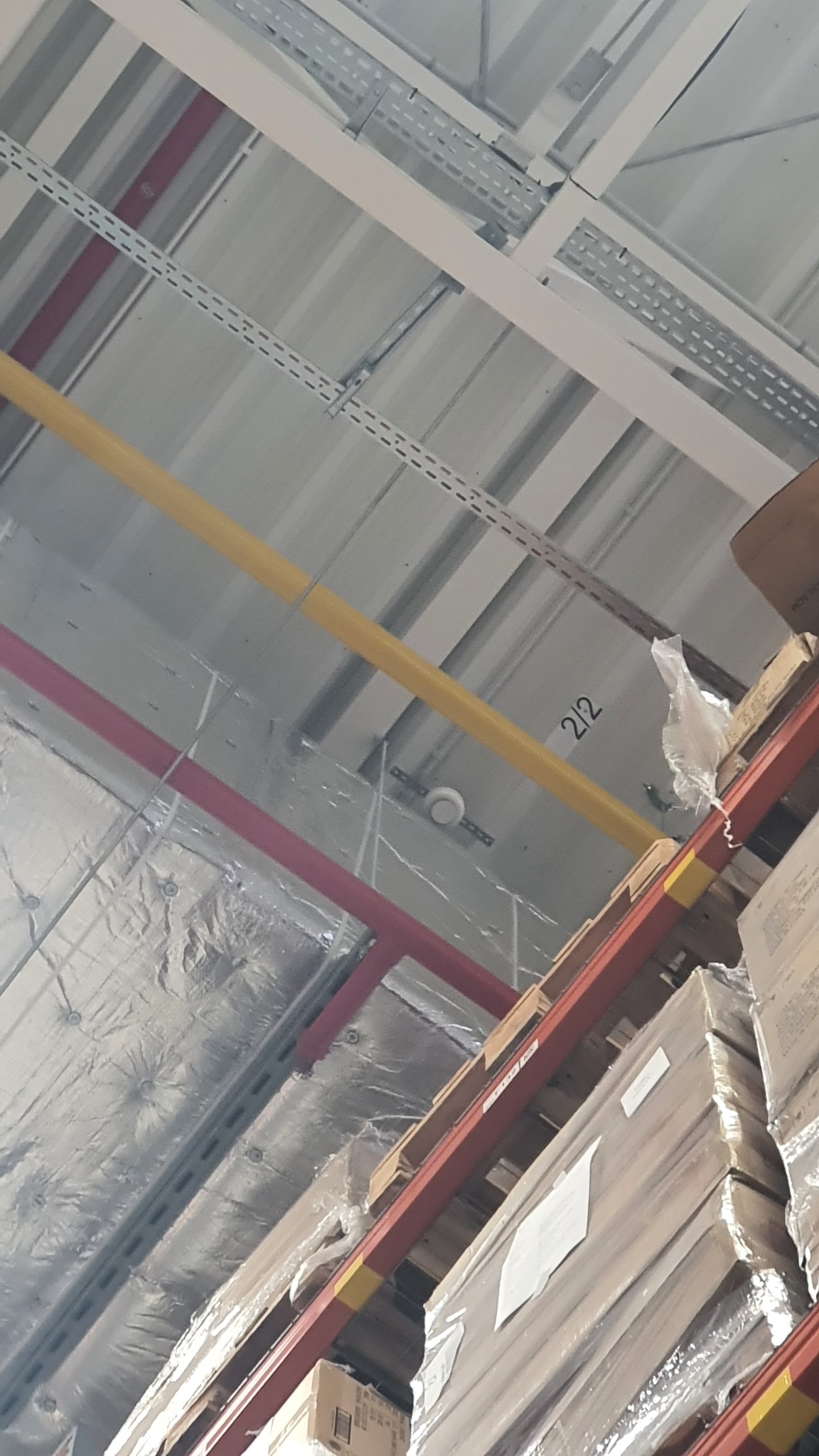 Widok w górę na sufit magazynu z widocznymi instalacjami elektrycznymi, rurami w kolorze żółtym i czerwonym, oraz systemem mocowań kablowych, a także elementami konstrukcyjnymi regałów magazynowych...