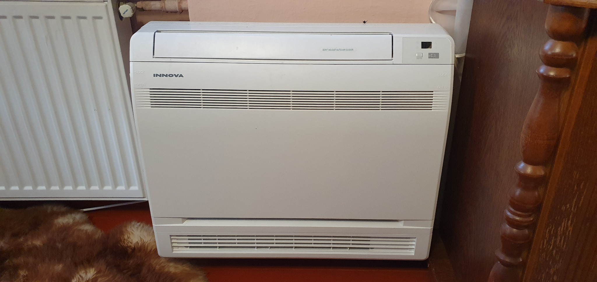Kompaktowa, biała klimatyzacja podłogowa marki Innova zainstalowana w pomieszczeniu z brązową podłogą, obok grzejnika i drewnianego mebla.