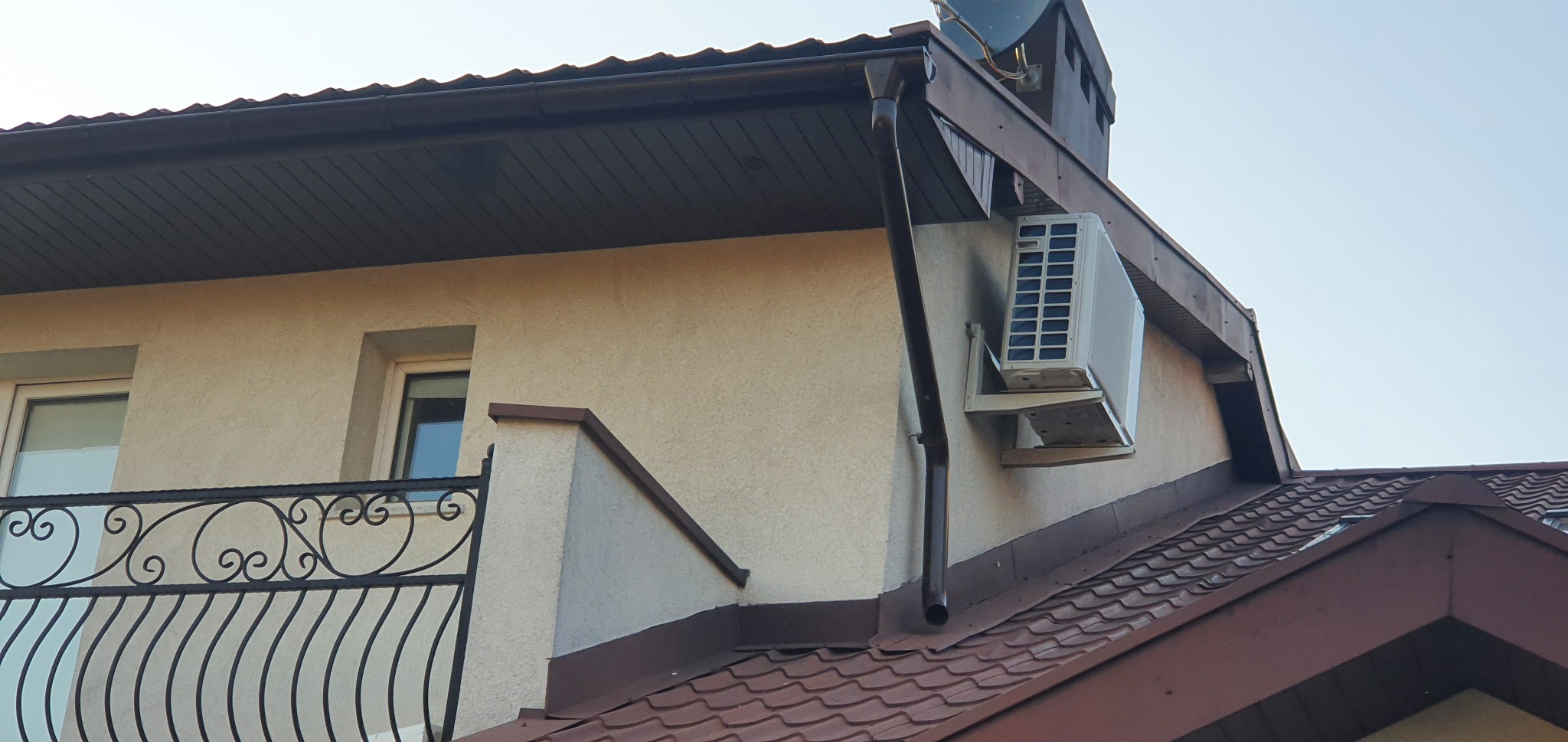 Zewnętrzna jednostka klimatyzacji zamontowana na ścianie domu z brązowym dachem, widoczny fragment balkonu z ozdobną balustradą i rynna odprowadzająca wodę.