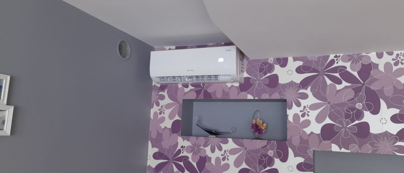 Biała jednostka klimatyzacyjna zamontowana pod sufitem w pokoju z szarą ścianą i ścianą w fioletowe kwiaty, widoczny okrągły nawiewnik wentylacyjny na szarej ścianie.