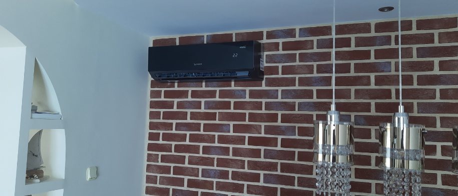 Czarna jednostka klimatyzacji zamontowana na ścianie z cegły dekoracyjnej, widoczny wyświetlacz temperatury i fragment ozdobnej lampy wiszącej.