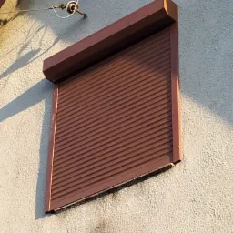 Ciemnobrązowa, częściowo opuszczona osłona okienna z poziomych listew na teksturowanej, jasnej ścianie zewnętrznej budynku, oświetlona z boku popołudniowym słońcem.