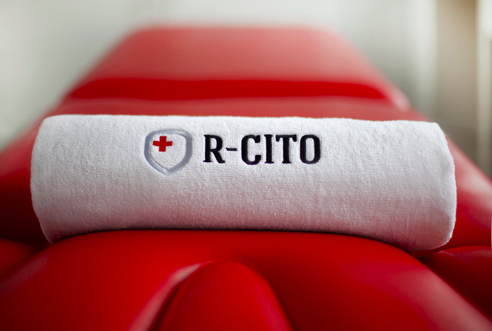 Czysty, biały ręcznik z logo firmy R-CITO, umieszczony na czerwonym, skórzanym stole do zabiegów.