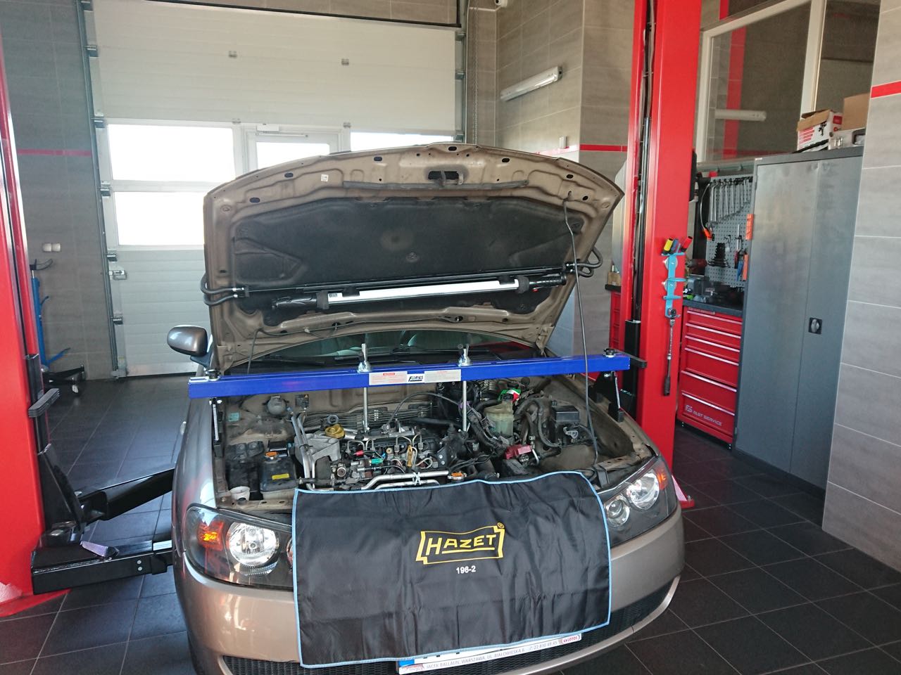 Samochód na podnośniku w warsztacie, z otwartą maską, zabezpieczony pokrowcem z logo Hazet, widoczne narzędzia i szafki warsztatowe.