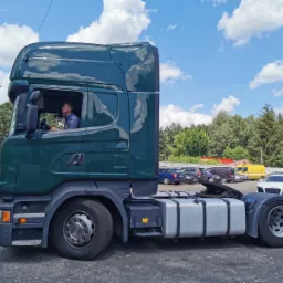 Zielony ciągnik siodłowy Scania z kierowcą w kabinie na parkingu w Warszawie, widok z boku na tle błękitnego nieba z chmurami.