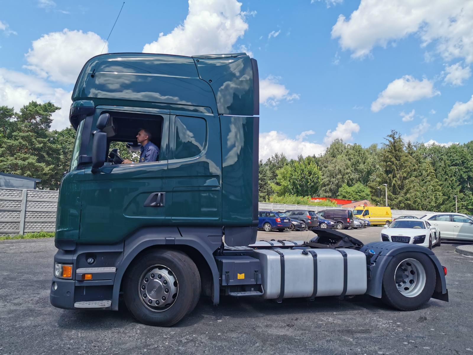 Zielony ciągnik siodłowy Scania z kierowcą w kabinie na parkingu w Warszawie, widok z boku na tle błękitnego nieba z chmurami.