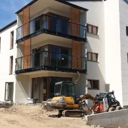 Nowoczesny budynek wielorodzinny z balkonami z drewnianymi przesłonami, w trakcie niwelacji terenu z użyciem koparek, widoczne przygotowania do zagospodarowania terenu.