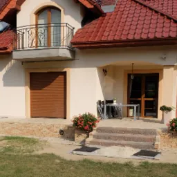 Elewacja domu jednorodzinnego z balkonem z ozdobną balustradą, tarasem wyłożonym jasnymi płytami, schodami wejściowymi z kamienia i rabatami kwiatowymi po bokach.