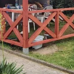 Fragment konstrukcji altany z krzyżowymi elementami z drewna pomalowanego na czerwono-brązowy kolor, widoczne białe krzesła i stół wewnątrz, roślina w doniczce na pniu drzewa na trawniku.