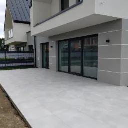 Nowoczesny taras wyłożony jasnoszarymi płytami kwadratowymi, przylegający do budynku z dużymi, przesuwnymi oknami i szarą elewacją. Widoczny fragment ogrodu i ogrodzenia w tle.