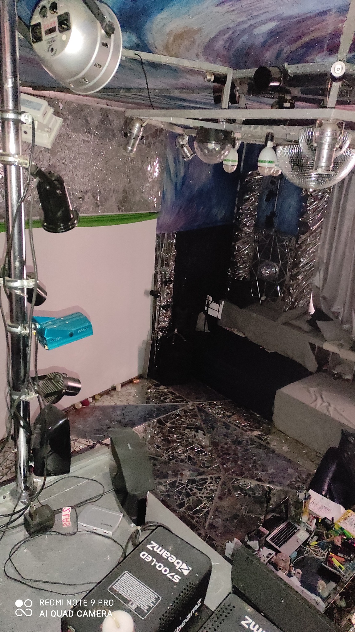 Scena z oświetleniem dyskotekowym: kule lustrzane, reflektory, efekty świetlne, srebrna folia dekoracyjna na ścianach i suficie, widoczne kable i sprzęt nagłośnieniowy, biała ściana w tle.