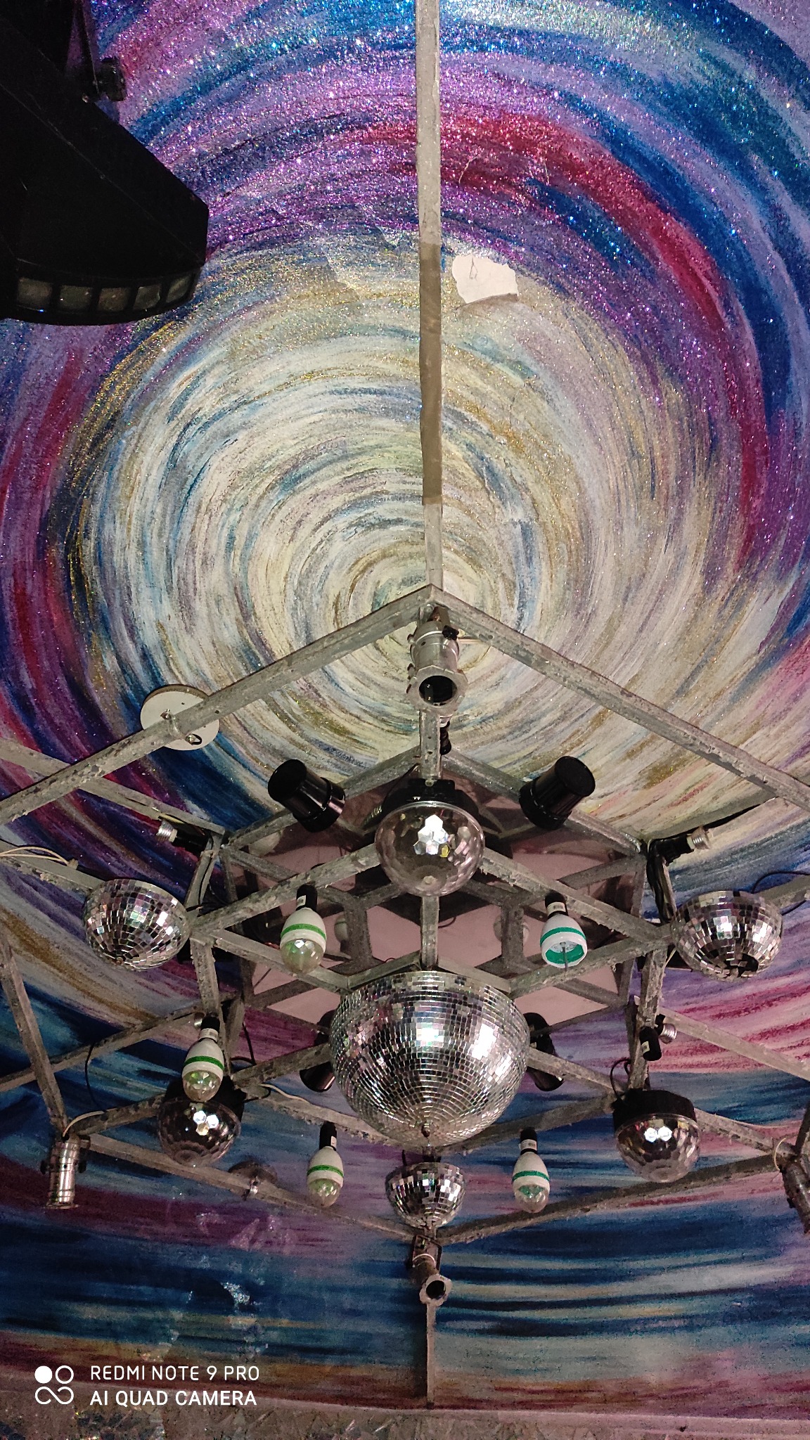 Sufit klubu nocnego z ręcznie malowanym wzorem galaktyki, metalowa konstrukcja z kulami dyskotekowymi i reflektorami, widok z dołu.