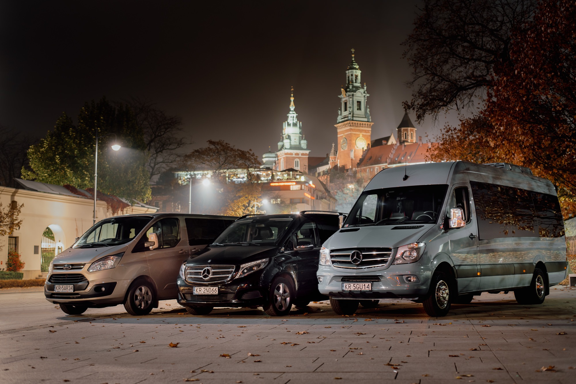 Trzy busy: Ford Transit, Mercedes V-klasa i Mercedes Sprinter, zaparkowane na tle nocnej panoramy Wawelu w Krakowie, oświetlone ulicznymi latarniami.