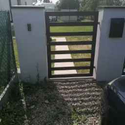 Nowoczesna brama wjazdowa z poziomymi, czarnymi szczeblami, osadzona w białych, murowanych słupkach z domofonem i skrzynką na listy, widok z zewnątrz na kamienisty podjazd i fragment samochodu.