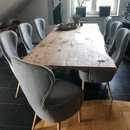 Stół z litego drewna o nieregularnych krawędziach, otoczony szarymi krzesłami na drewnianych nogach, ustawiony w pomieszczeniu z czarną podłogą i oknem z roślinami.