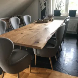 Stół z blatem z litego drewna o nieregularnej krawędzi, otoczony szarymi krzesłami na drewnianych nogach, w tle okno i dekoracje.