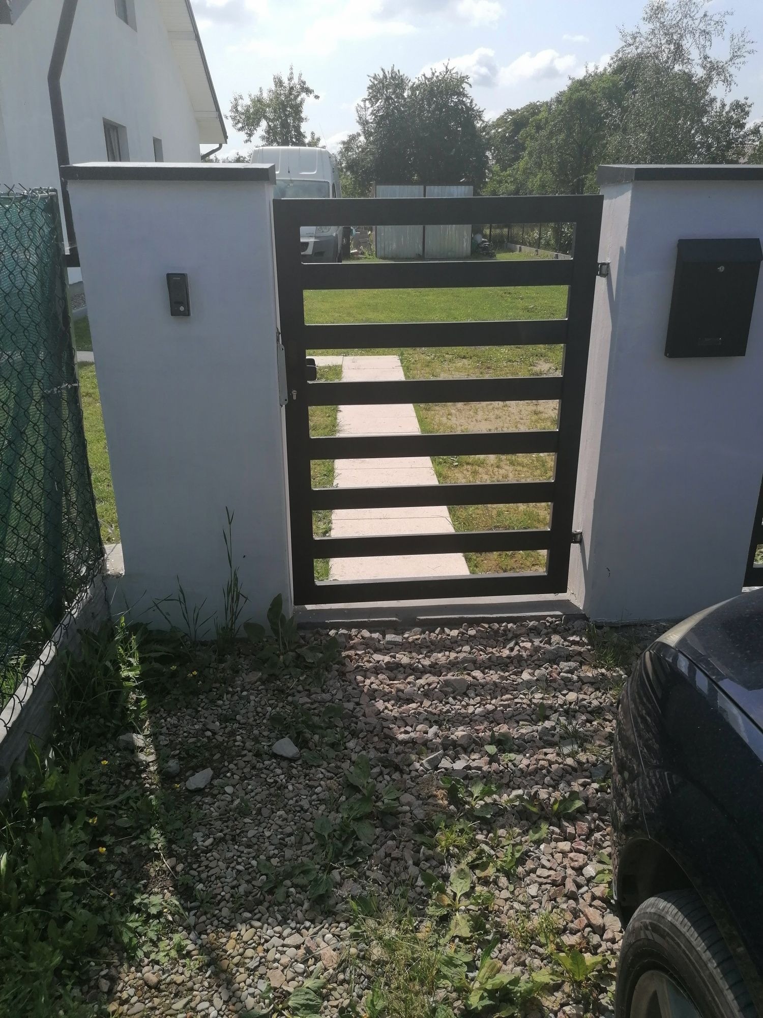 Nowoczesna brama wjazdowa z poziomymi, czarnymi szczeblami, osadzona w białych, murowanych słupkach z domofonem i skrzynką na listy, widok z zewnątrz na kamienisty podjazd i fragment samochodu.