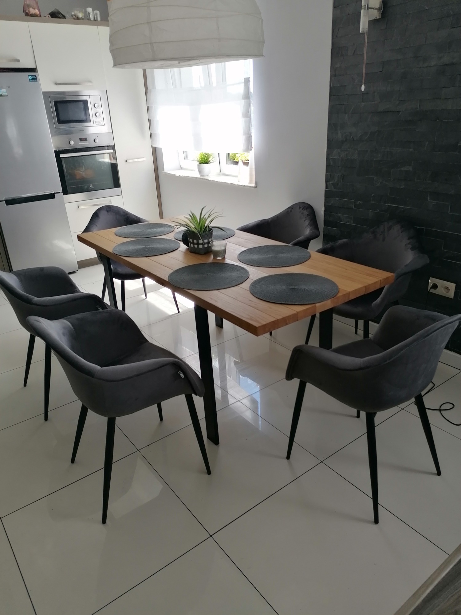 Stół z jasnego drewna na czarnych nogach otoczony sześcioma szarymi krzesłami z welurowym obiciem i czarnymi nogami, nakryty okrągłymi, szarymi podkładkami, na tle białej kuchni z lodówką...
