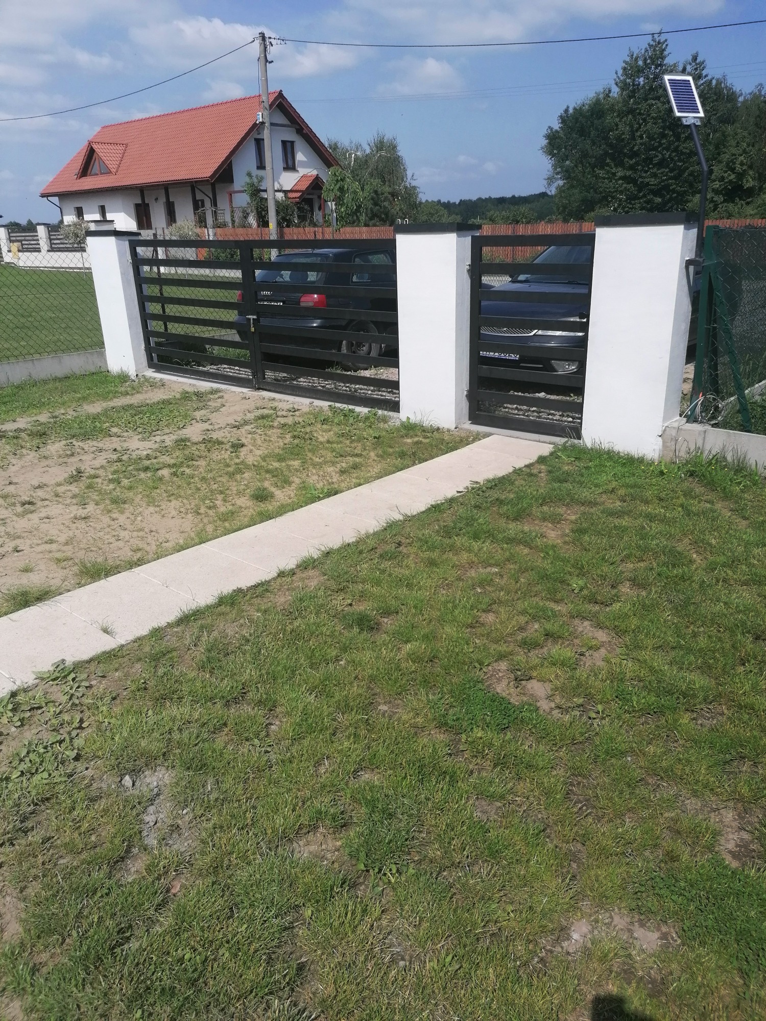 Nowoczesna, czarna brama wjazdowa z poziomymi szczeblami, osadzona w białych, murowanych słupkach. Widoczny fragment posesji z samochodem w tle oraz panel solarny zamontowany na słupku obok bramy.