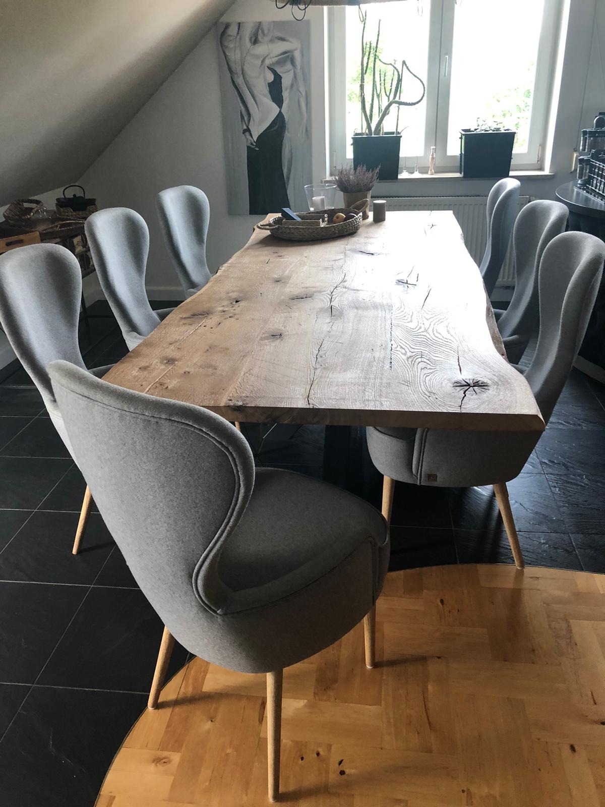 Stół z litego drewna o nieregularnych krawędziach, otoczony szarymi krzesłami na drewnianych nogach, ustawiony w pomieszczeniu z czarną podłogą i oknem z roślinami.