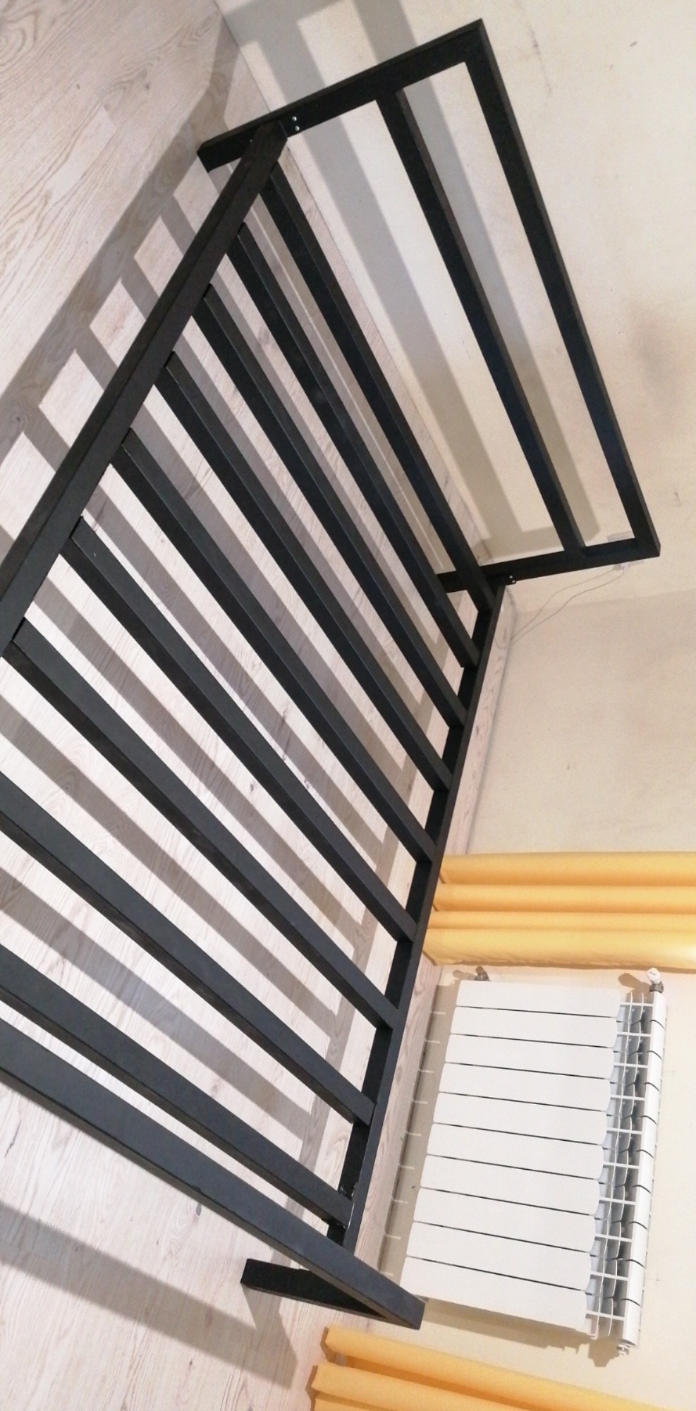 Częściowo zmontowana, czarna, metalowa konstrukcja z prętów, prawdopodobnie element balustrady lub ogrodzenia, leżąca na podłodze w jasnym pomieszczeniu, widoczny grzejnik i zabezpieczenia rur.