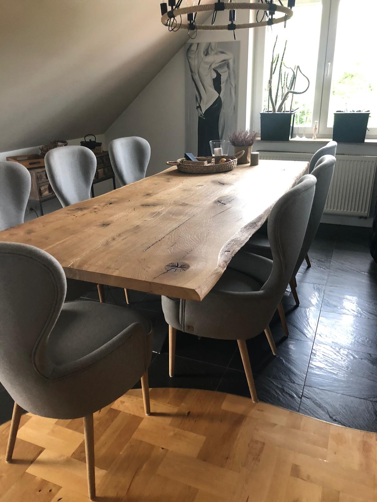 Stół z blatem z litego drewna o nieregularnej krawędzi, otoczony szarymi krzesłami na drewnianych nogach, w tle okno i dekoracje.