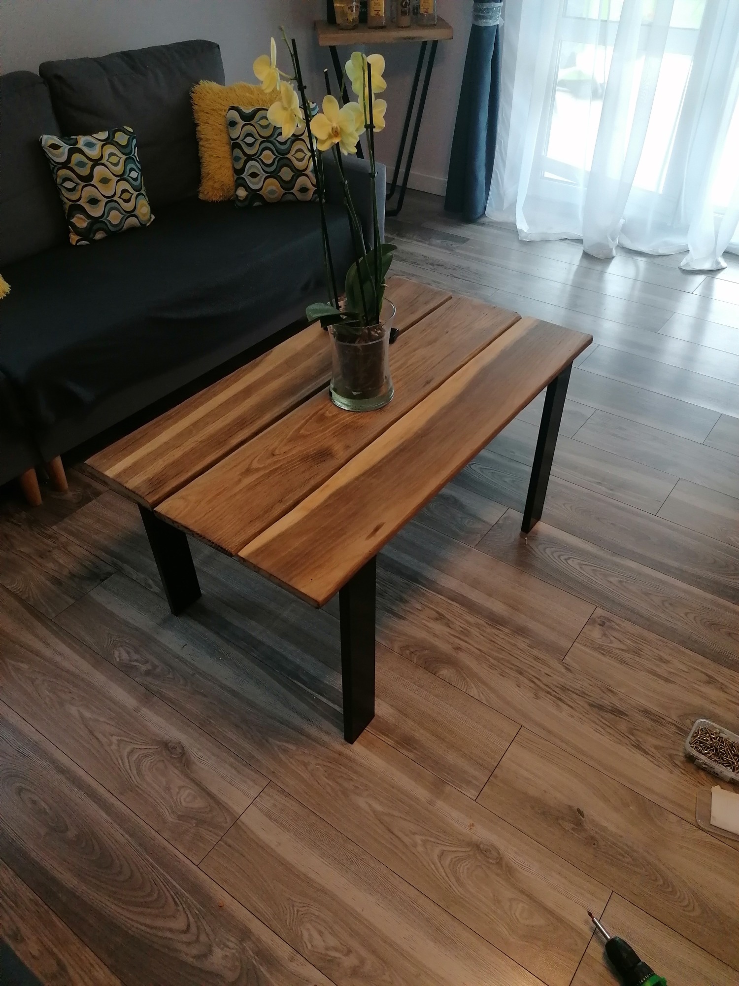 Stolik kawowy z blatem z desek o ciepłym odcieniu drewna i czarnymi, metalowymi nogami, na podłodze panele imitujące drewno, w tle szara sofa z poduszkami i żółta orchidea w szklanym wazonie...