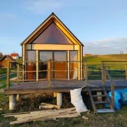 Nowoczesny, drewniany domek letniskowy z dużym przeszkleniem w szczycie, posadowiony na betonowych słupach z drewnianym tarasem, widoczne ślady budowy: worki z odpadami i resztki drewna.