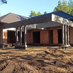 LIVBUD Marcin Skrzat - Budowa domu z czerwonej cegły w stanie surowym otwartym, widoczne betonowe słupy i zadaszenie. W tle drzewa i błękitne niebo.