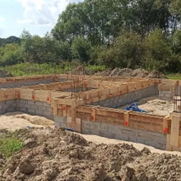 Wykopane fundamenty domu z szalunkiem i zbrojeniami, przygotowane do wylania betonu, widok z góry na tle zieleni.