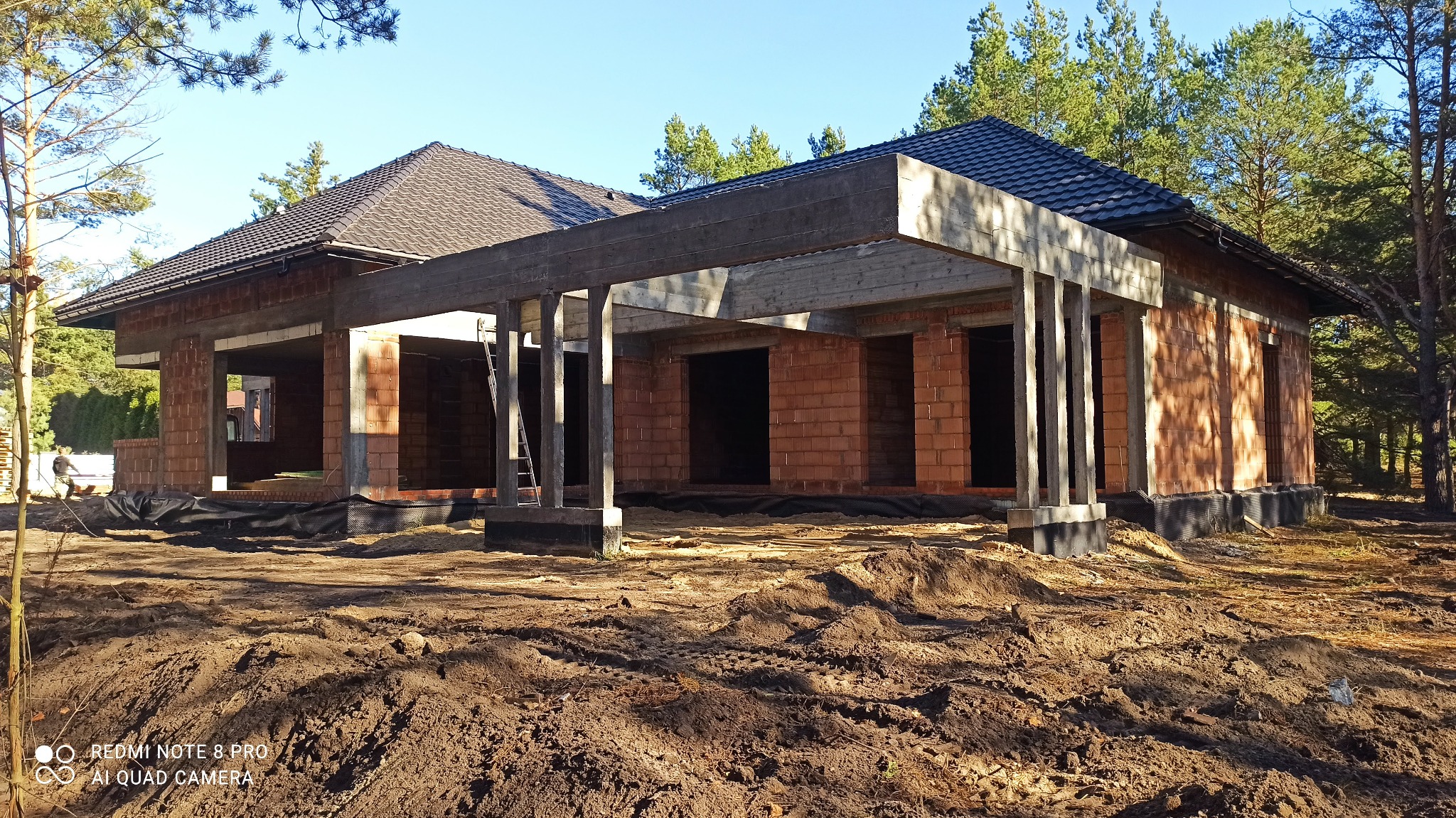 Budowa domu z czerwonej cegły w stanie surowym otwartym, widoczne betonowe słupy i zadaszenie. W tle drzewa i błękitne niebo.
