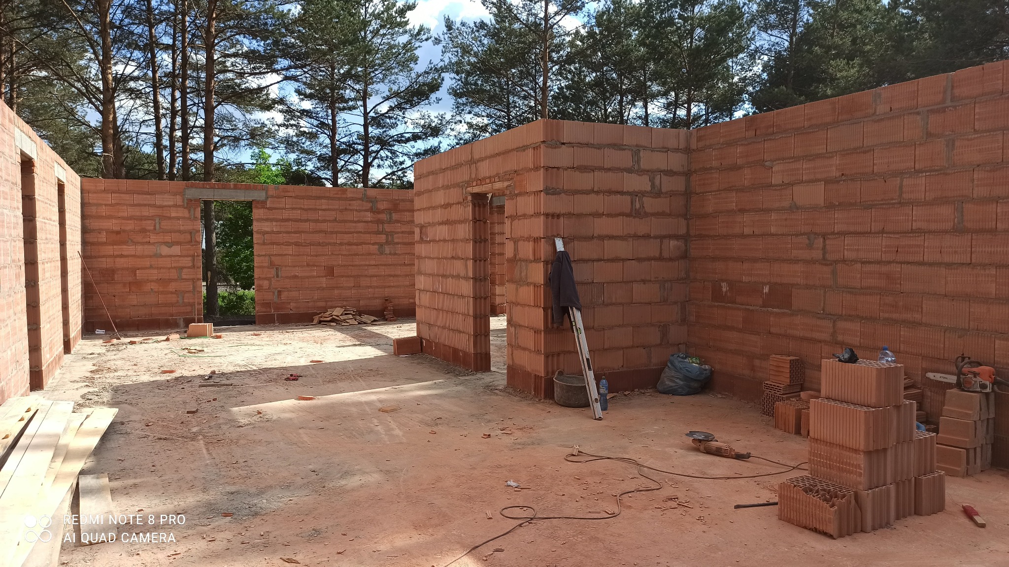 Wnętrze domu w trakcie budowy, ściany z czerwonej cegły, widoczne otwory drzwiowe, w tle drzewa. Na podłodze leżą cegły, narzędzia i drabina.