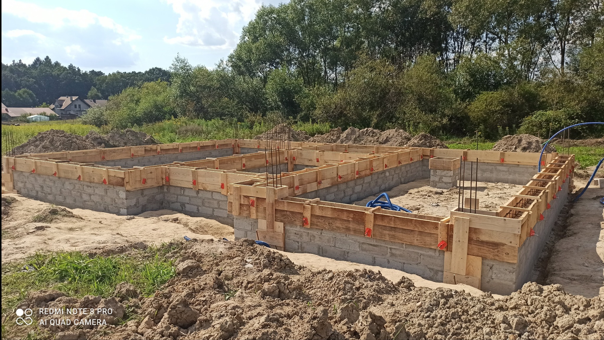 Wykopane fundamenty domu z szalunkiem i zbrojeniami, przygotowane do wylania betonu, widok z góry na tle zieleni.