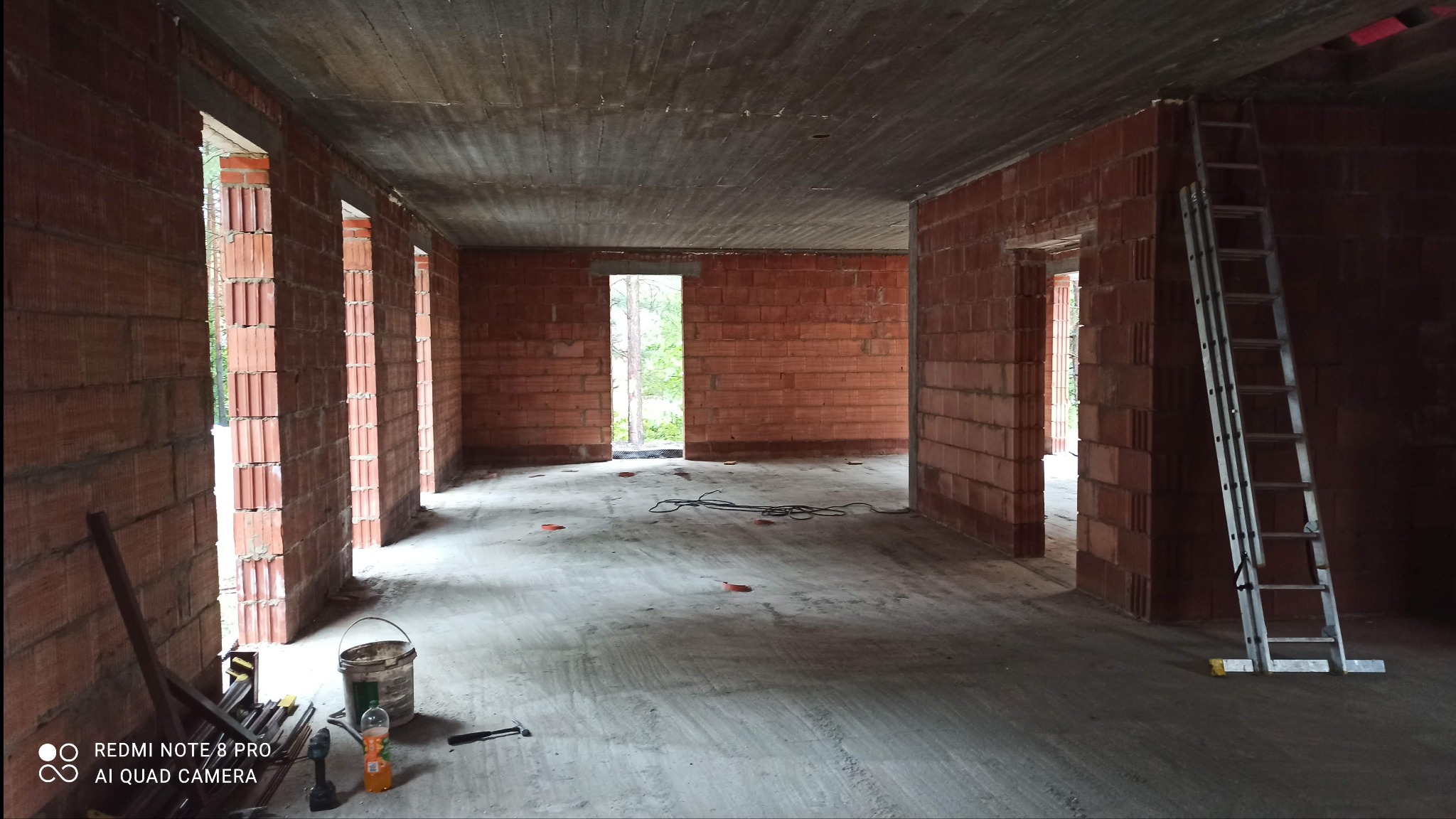 Wnętrze domu w trakcie budowy, widoczne ściany z czerwonej cegły, surowy betonowy strop i otwory okienne. Na podłodze leżą kable i narzędzia, a przy ścianie stoi drabina.