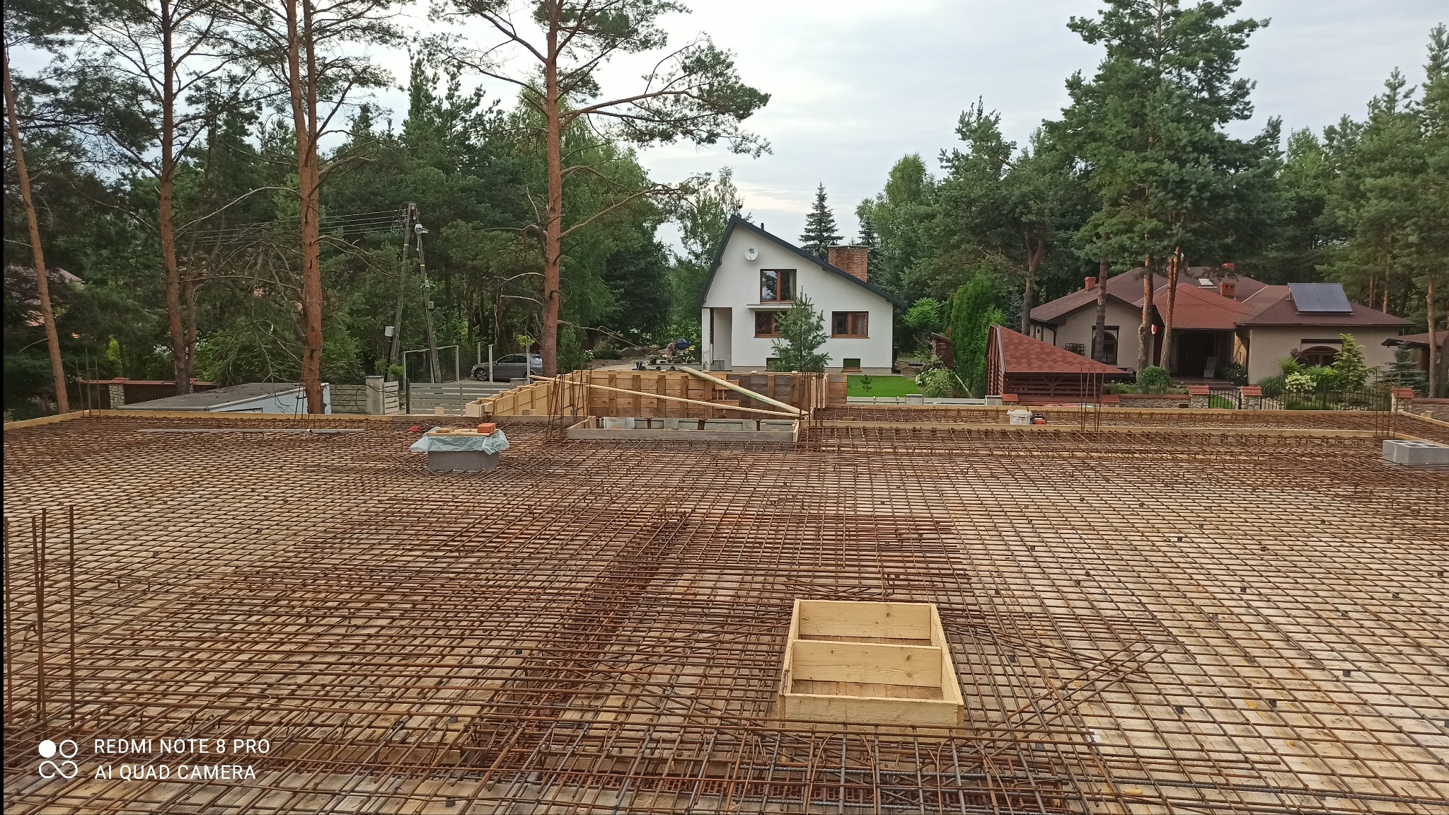 Zbrojenie płyty fundamentowej domu jednorodzinnego, widok z góry na siatkę z prętów i szalunki. W tle domy i drzewa.