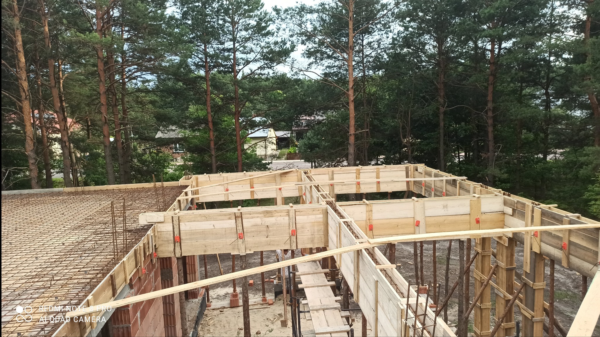 Wysoka perspektywa na szalunek stropu z widocznym zbrojeniem, tle drzewa i fragment budynku. Detal konstrukcji z drewnianych belek i metalowych podpór.