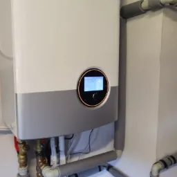 Nowoczesny, biało-szary piec z dotykowym panelem sterowania, widoczne rury z izolacją termiczną i zawory, zamontowany na białej ścianie w pomieszczeniu technicznym.