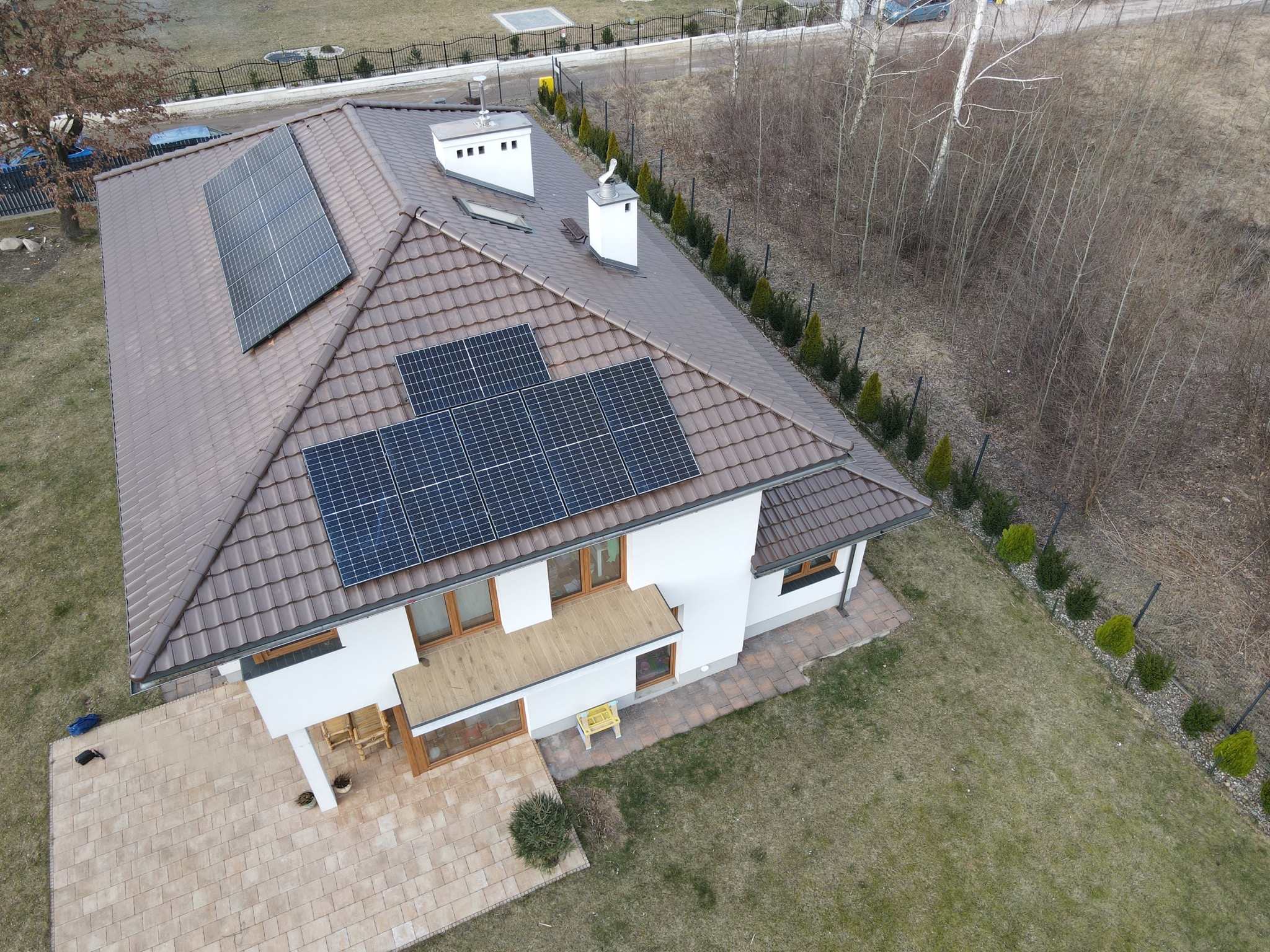 Widok z góry na dom jednorodzinny z brązowym dachem pokrytym panelami słonecznymi, białą elewacją i zadbanym ogrodem, z widocznym tarasem i elementami małej architektury.