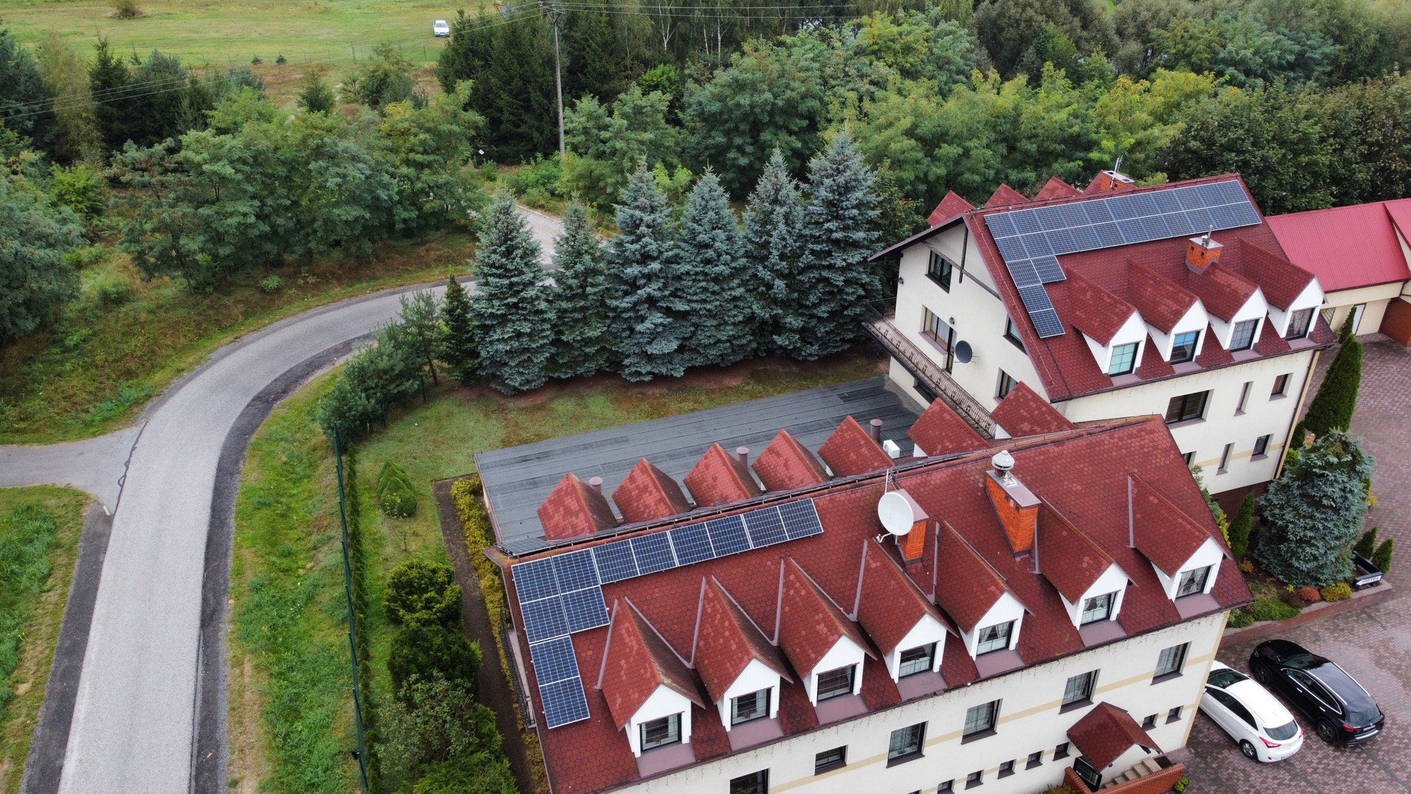 Widok z góry na dwa budynki z czerwonymi dachami, na których zamontowano panele słoneczne; w tle droga, drzewa iglaste i liściaste, a na parkingu zaparkowane dwa samochody.