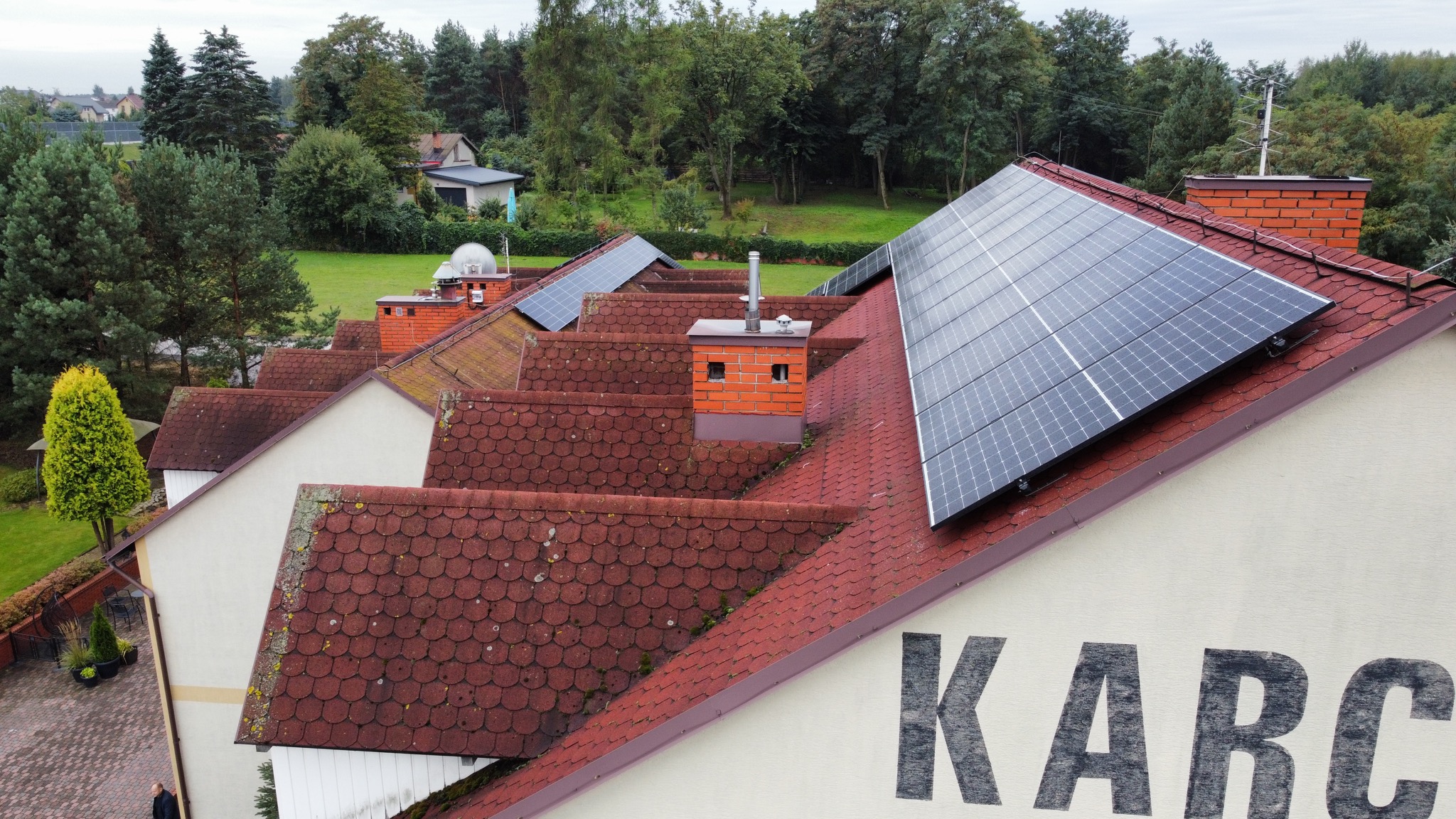 Widok z góry na dom z panelami słonecznymi na dachu pokrytym czerwoną dachówką, ceglane kominy i zielone drzewa w tle.