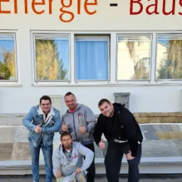 Czterech uśmiechniętych mężczyzn, prawdopodobnie ekipa budowlana, pozuje przed budynkiem z napisem 'Energie - Baus', pokazując kciuki w górę; widoczne próbki materiałów elewacyjnych.