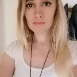 Portret kobiety z blond włosami w Łodzi, ubrana w białą koszulkę i długi naszyjnik, pozująca na tle jasnej ściany.