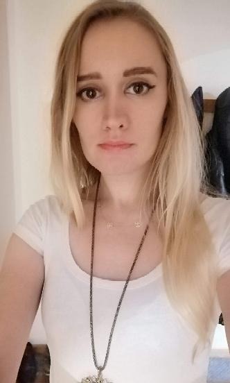 Portret kobiety z blond włosami w Łodzi, ubrana w białą koszulkę i długi naszyjnik, pozująca na tle jasnej ściany.