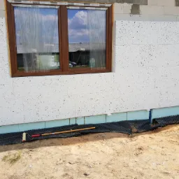 Ocieplona ściana budynku z oknem, zabezpieczona folią budowlaną, z widocznym wałkiem malarskim i długim uchwytem na ziemi.