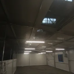 Wnętrze hali produkcyjnej lub magazynowej z widoczną konstrukcją stropu, oświetleniem jarzeniowym i oknem w ścianie, na podłodze widoczne ślady użytkowania.